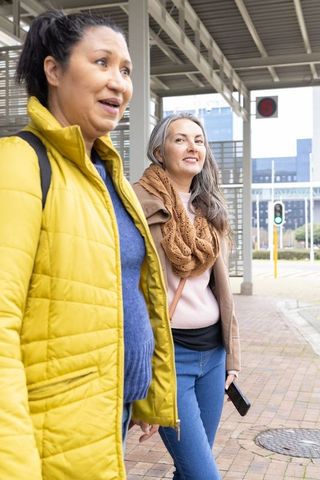Mature women walking at tram stop wearing yellow jacket and cozy scarf holding smartphone