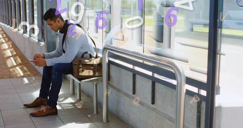 Commuter scrolling smartphone while waiting on transit bench with messenger bag, sunlit