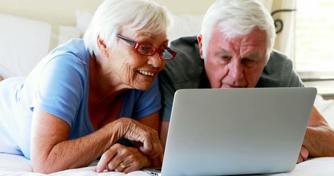 Senior couple lying on bed laughing while using laptop