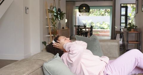 African american woman reclining on sofa wearing white headphones in pink loungewear