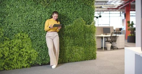 Young professional leaning on green wall using tablet in modern open-plan office