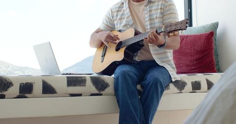 Mature man playing acoustic guitar on sunlit window seat with laptop and colorful cushions