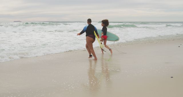 Happy african american female friends running into the sea holding surfboards. healthy outdoor leisure time by the sea.