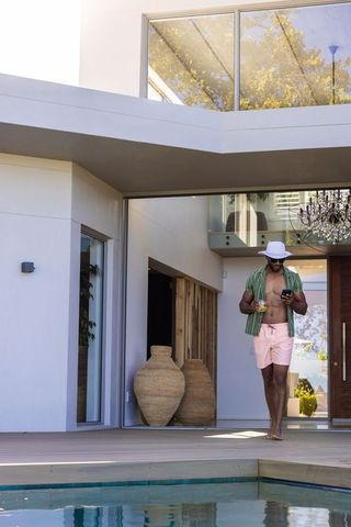 Mature african man walking on pool deck checking smartphone holding drink at modern villa