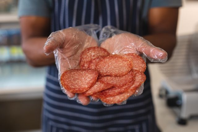 Midsection of african american young male owner holding salami while standing in cafe. unaltered, hygiene, gloves, cafeteria, barista, occupation, food and small business concept.