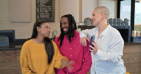 Three friends chatting at coffee shop counter sharing smartphone and laughing together