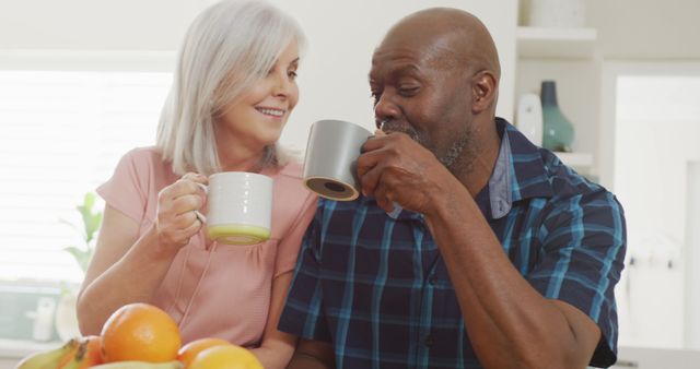 A senior couple is enjoying their morning coffee together while smiling warmly at each other. The couple, dressed in casual attire, appears to be engaging in relaxed conversation in the kitchen. The table in front of them has a small assortment of fresh fruit, adding a healthy and serene atmosphere to the scene. This image is ideal for promoting senior lifestyle products, illustrating the importance of companionship, and highlighting indoor family activities.