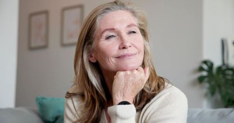 Pensive Senior Woman Relaxing on Sofa with Wristwatch in Minimalistic Living Room