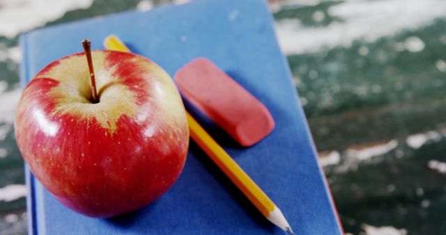 A red apple sits atop a blue book with a pencil and eraser against a backdrop of a used chalkboard, with copy space. Symbolizing traditional education, the apple represents a gift for the teacher or a healthy snack for a student.