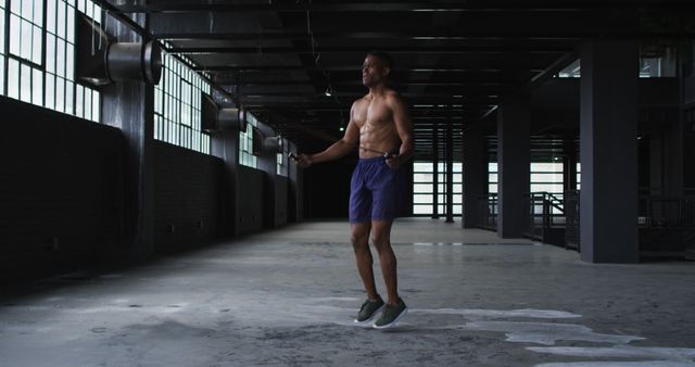 Shirtless man jumping rope in a spacious industrial-style gym, emphasizing fitness and health. Great for promotional materials on physical training, workout routines, motivational fitness campaigns, or athletic apparel advertisements.