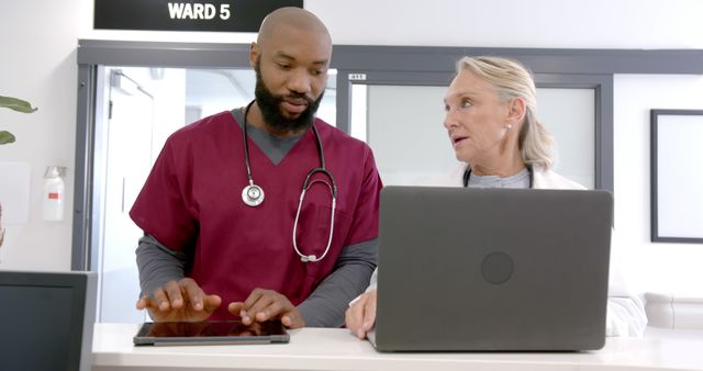 Medical professionals are discussing patient data in a hospital ward. One professional is in red scrubs holding a tablet, while another in white coat works on a laptop. This image is useful for illustrating healthcare teamwork, modern medical practices, or hospital environments.