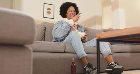 African american man relaxing on sofa eating popcorn pointing and smiling at tv