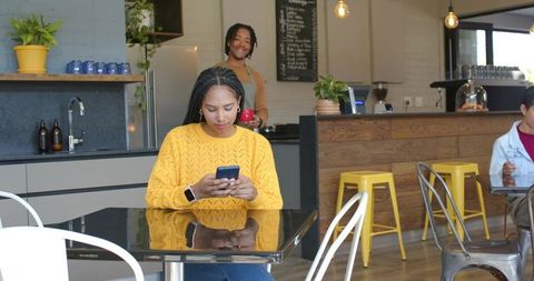African american woman using smartphone at modern cafe in yellow sweater and smartwatch