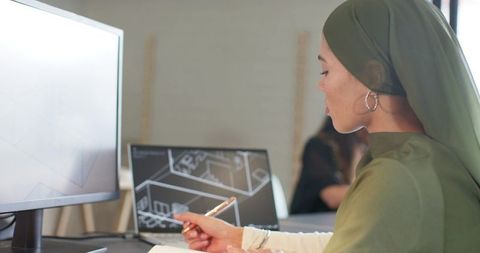 Indian woman in green hijab pointing at laptop with architectural cad blueprints in modern office wo