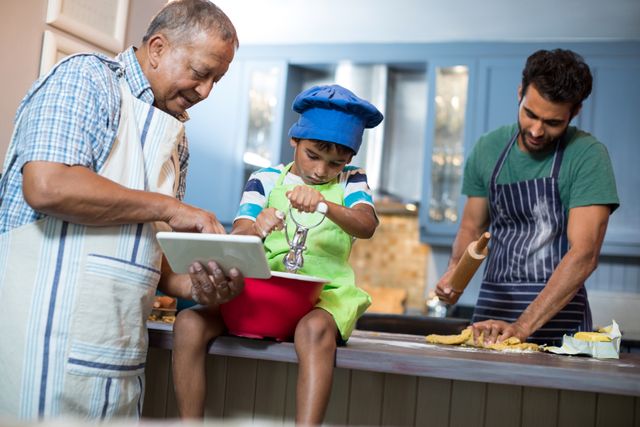 Grandfather showing a tablet to his grandson while the boy is using a kitchen tool, and the father is rolling dough in the background. This image can be used to depict family bonding, multigenerational activities, and the joy of cooking together. Ideal for advertisements, blogs, and articles related to family life, cooking, and technology in the kitchen.