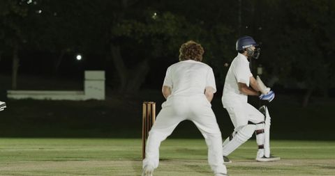 Night cricket match with batsman playing defensive shot under floodlights on grass pitch