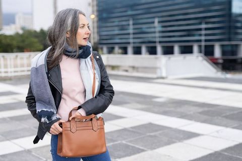 Mature woman standing city plaza holding leather handbag wearing leather jacket and scarf