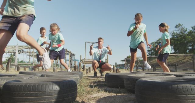 Group of children engaging in an outdoor obstacle course under the guidance of a coach. They navigate through tires, showcasing their agility and teamwork. Useful for campaigns promoting physical activity, teamwork in schools, children's fitness, outdoor events, or summer camps.