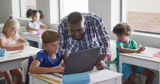 Image of happy african american male teacher helping caucasian boy with laptop. primary school education and teaching profession.