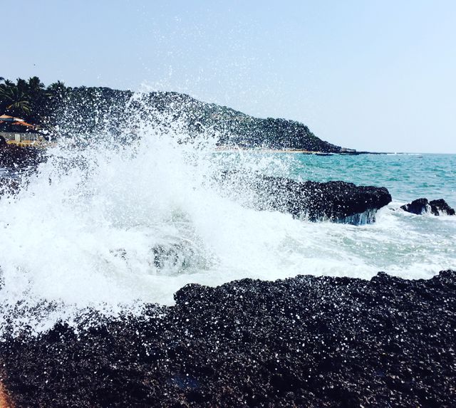 Waves crashing against black rocks while splashing water under clear sky. Coastal view showcasing strength of ocean waves meeting land. Suitable for use in projects related to beaches, marine biology, travel blogs, vacation brochures, and nature conservation content.