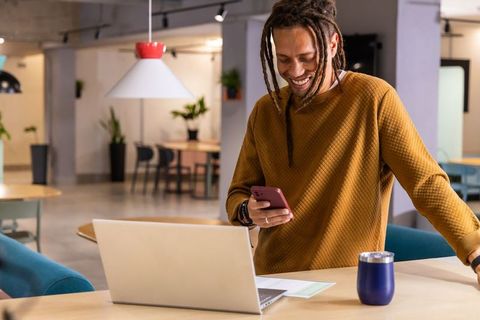 African American man standing at coworking desk smiling and using smartphone with laptop