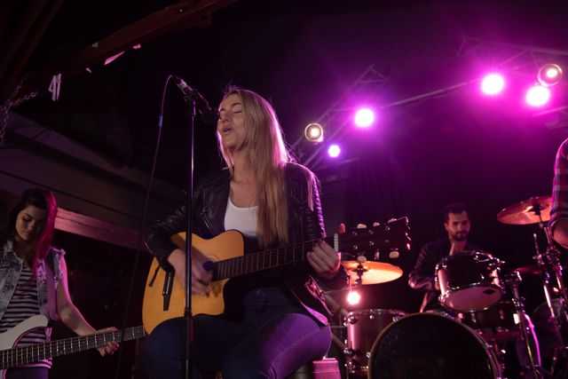 Caucasian woman sitting and playing acoustic guitar, female guitarist, male drummer and band equipment on stage at a music venue during concert in the background. Entertainment fun music.