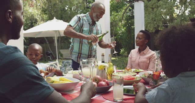 Happy african american family sitting at table in garden, eating dinner and drinking wine. Lifestyle, domestic life, family, and togetherness.