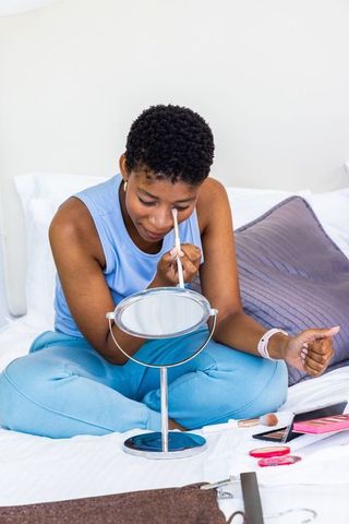African woman applying eye makeup on bed using hand mirror for self-care routine