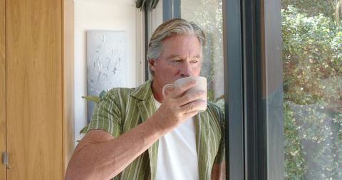 Senior man standing by sliding glass door holding mug gazing outside in soft daylight
