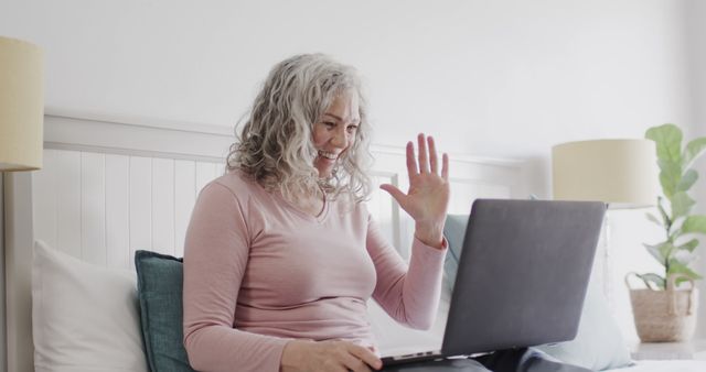 Senior woman with grey hair happily video chatting on a laptop from her bed at home. Can be used to depict technology use among elderly, remote communication, online interactions, family connections, or social engagement activities. Suitable for advertisements about communication devices, social apps, or articles on elder tech usage, staying connected, and emotional well-being.