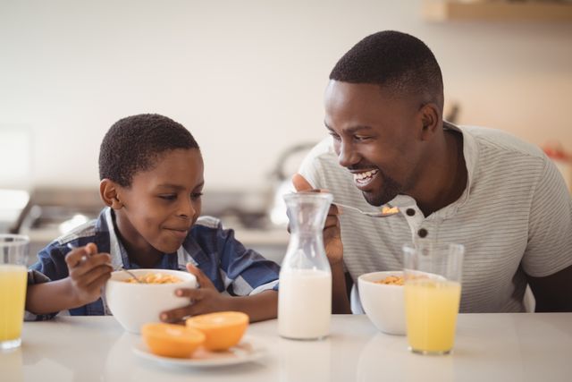 Father and son sharing a joyful breakfast in a modern kitchen. Both are eating cereal with milk, accompanied by glasses of orange juice. Ideal for use in family-oriented advertisements, parenting blogs, and healthy lifestyle promotions.