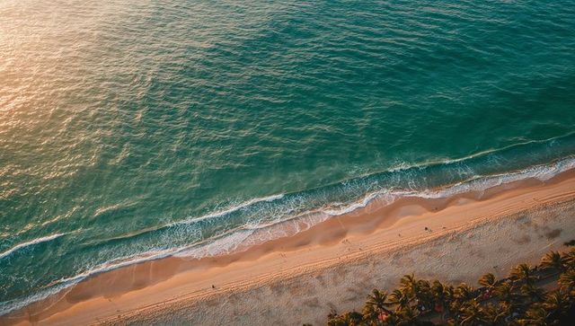 Sunlit turquoise ocean rolling onto pristine sandy beach from drone aerial view at sunset