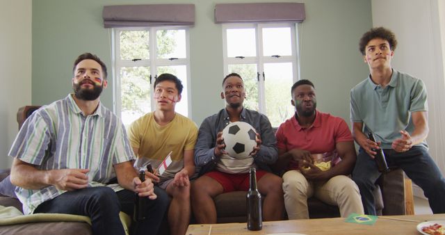 A group of diverse male friends is sitting on a couch in a living room watching a soccer game on TV. A man in the center is holding a soccer ball, and they all appear to be cheering. Some have their faces painted to support their team, and there are snacks and drinks on the table. This image can be used for advertising sports events, promoting social activities, or illustrating moments of camaraderie and friendship.