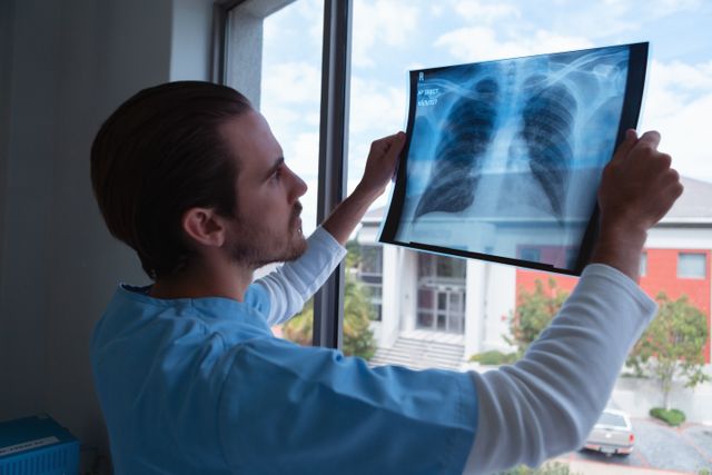 Male surgeon in blue scrubs examining a chest x-ray near a window in a hospital. Useful for content related to healthcare, medical diagnostics, radiology, and professional medical services.