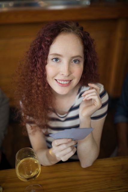 Smiling woman with curly red hair holding cards while sitting at a wooden table in a bar. She is wearing a striped shirt and appears to be enjoying a casual and relaxed moment. A glass of white wine is on the table. This image can be used for themes related to leisure, socializing, relaxation, and casual gatherings.