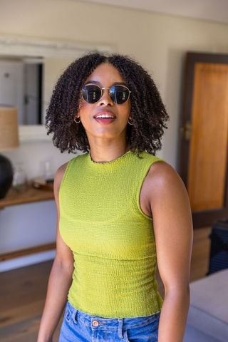 African american woman wearing sunglasses and lime green top standing in modern room