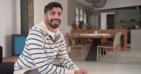 Indian man smiling sitting in bright modern open-plan living room wearing striped hoodie