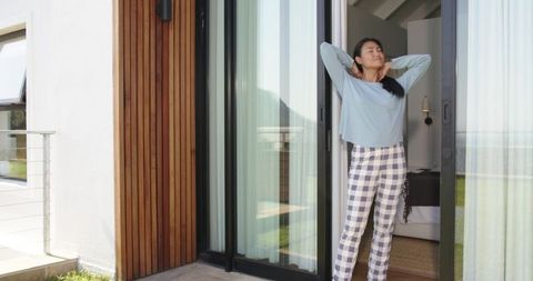 Mid-adult asian woman stretching in sliding door of modern home wearing checked pants
