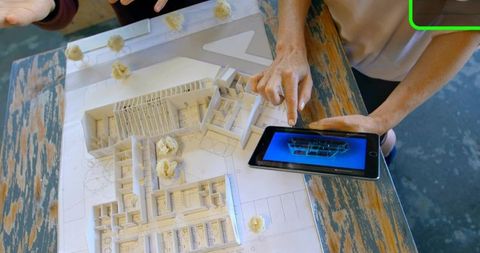 Designers in sleeveless tops collaborating over architectural model and tablet rendering
