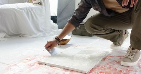 Indian man kneeling spreading tile adhesive with notched trowel during flooring renovation