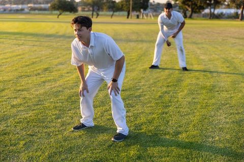 Two cricketers practicing close fielding at sunset on green pitch wearing white kit