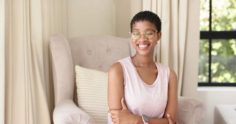 Smiling woman sitting on beige armchair wearing eyeglasses and smartwatch in sunlit living room