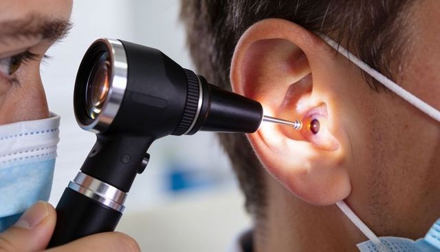 Masked clinician examining patient ear with otoscope and illuminated speculum close-up clinical view