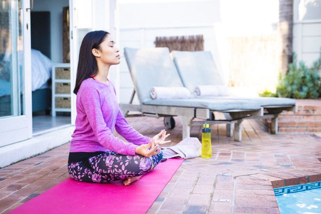 Biracial woman practicing yoga and meditating on a pink mat by the poolside. She is wearing a purple long-sleeve shirt and patterned leggings, focusing on relaxation and mindfulness. Ideal for use in articles or advertisements related to fitness, wellness, self-care, and healthy lifestyle.