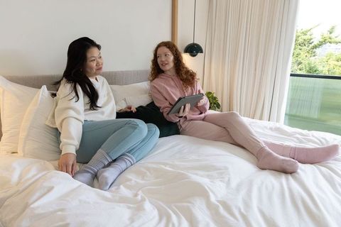 Diverse women relaxing on bed in bright minimalist bedroom holding tablet and chatting