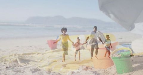 Family of four walking hand in hand on sunlit beach with colorful sand buckets and umbrella