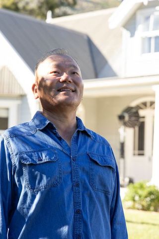 Mature asian man standing in front yard looking up under sunlight wearing denim shirt