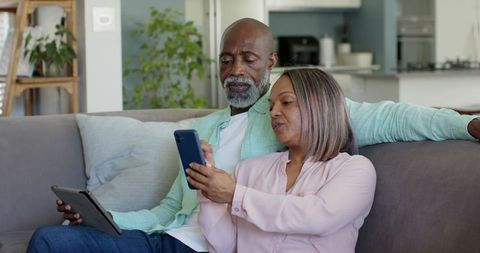 Senior couple relaxing on gray sofa using smartphone and tablet in modern living room
