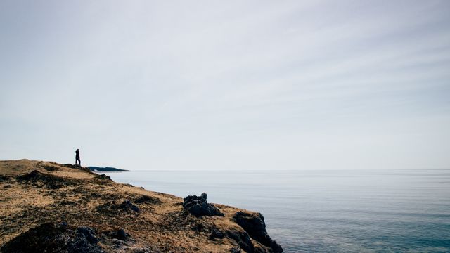This visually striking image captures a lone person standing on a rocky shore, overlooking a calm and expansive sea. The scene conveys feelings of solitude, contemplation, and serene natural beauty. Ideal for use in travel promotions, inspirational ads, mental health discussions, or blog posts about solitude and peaceful places.