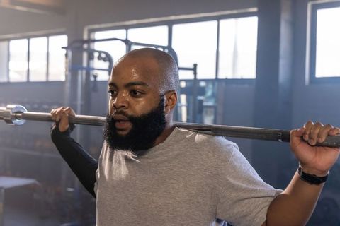 African american man performing barbell back squat in gym, sweating and focusing